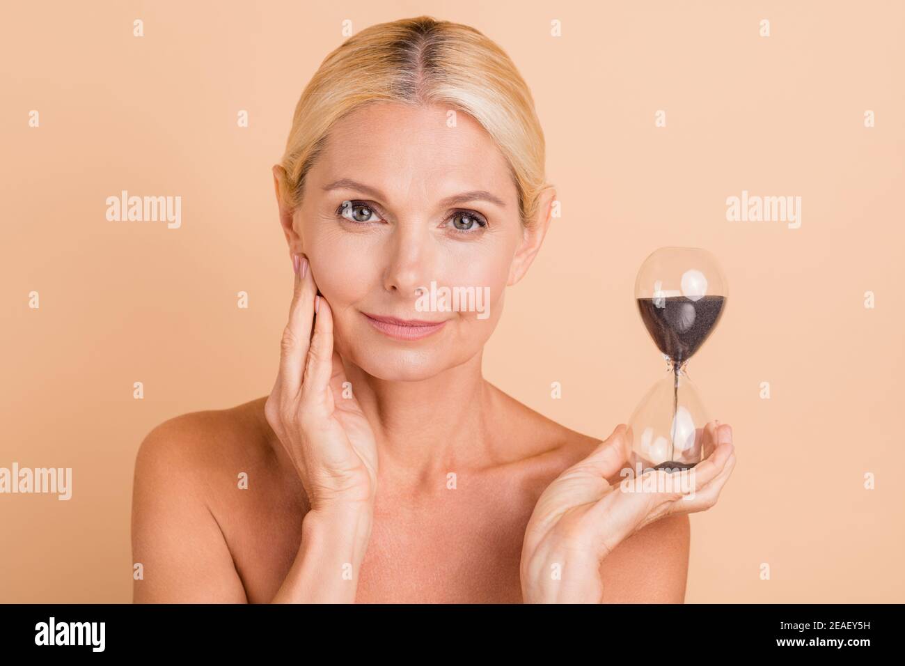 Portrait von Frau mittleren Alters halten Sand Stunde Glas berühren Ihre Haut Spa Salon Anti-Aging-Therapie isoliert beige Farbe Hintergrund Stockfoto