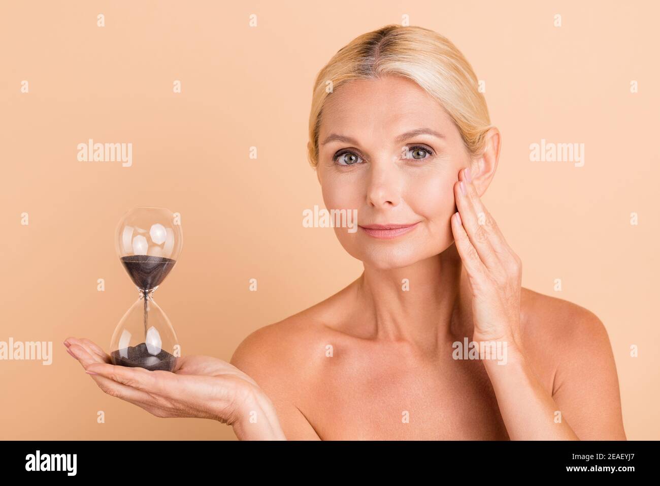 Portrait charmante Frau mittleren Alters halten Sandglas Uhr berühren Hände Wangen isoliert Pastellfarben Hintergrund Stockfoto
