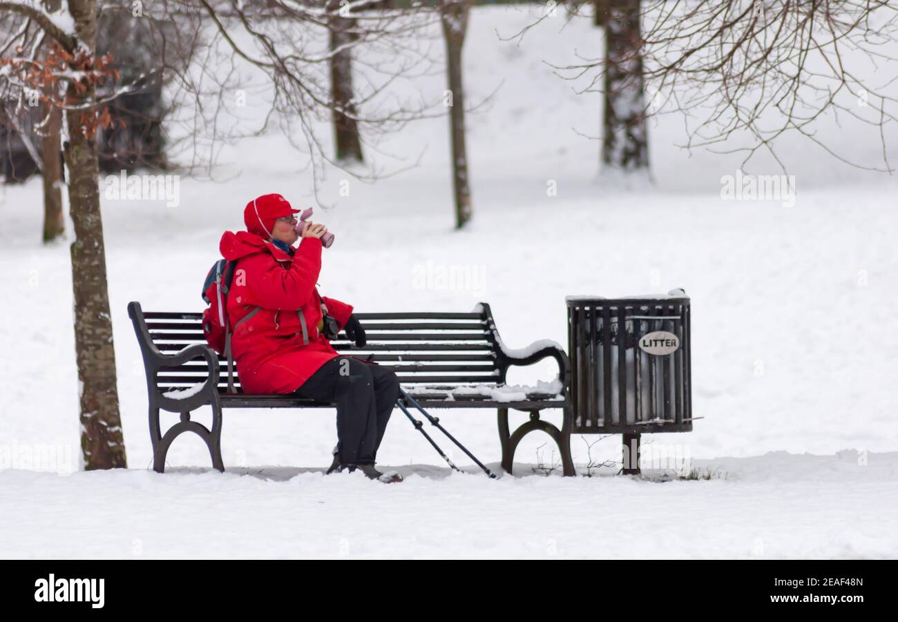 Glasgow, Schottland, Großbritannien. 9th. Februar 2021. UK Wetter: Eine Frau in rot gekleidet sitzt auf einer schneebedeckten Bank mit einem Drink in Queen's Park nach starkem Schneefall von Storm Darcy. Kredit: Skully/Alamy Live Nachrichten Stockfoto