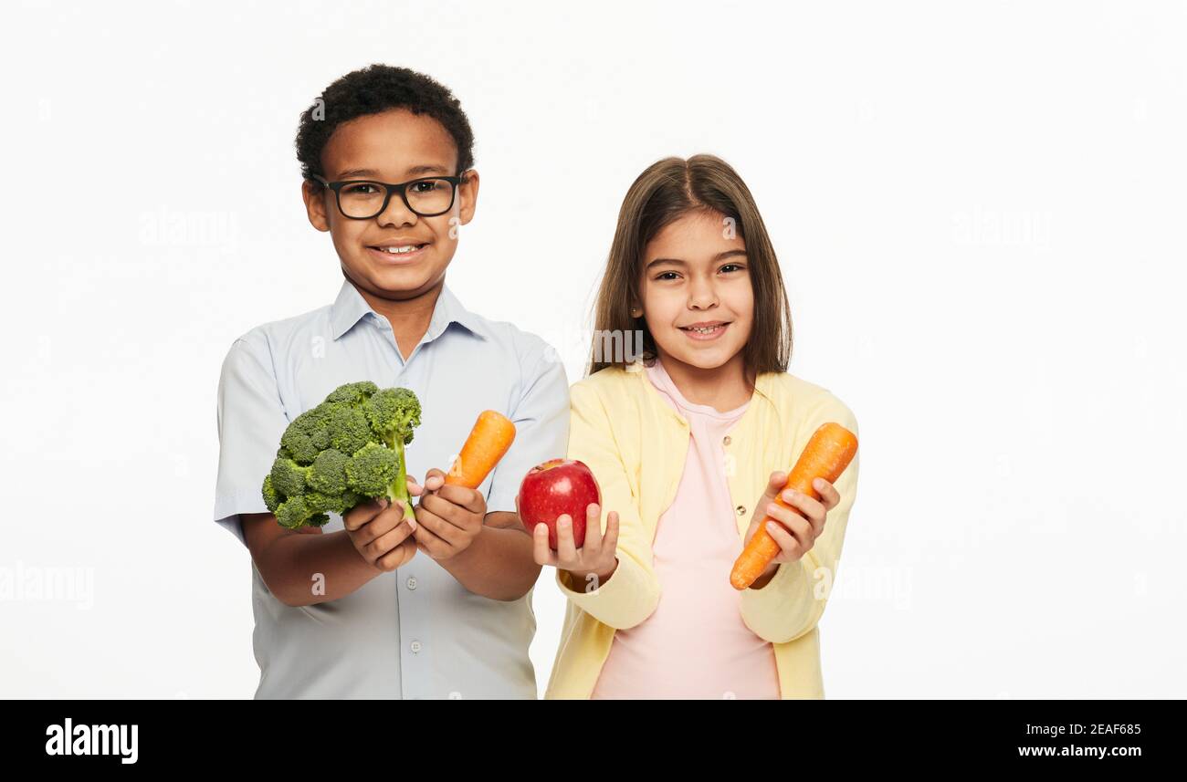 Latino Mädchen und afroamerikanischen Jungen halten Brokkoli, Karotten und einen Apfel. Vorteile von Gemüse und Obst für Kinder Stockfoto