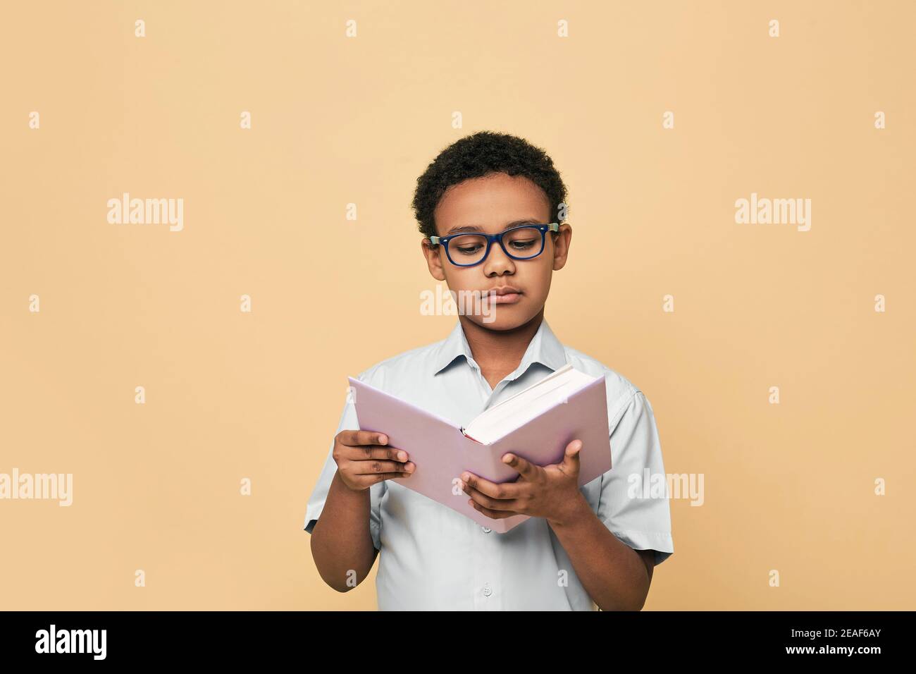 Afroamerikanischer kleiner Junge mit Brille hält Buch, das nachdenklich auf Schulbuch aussieht, auf beigem Hintergrund Stockfoto