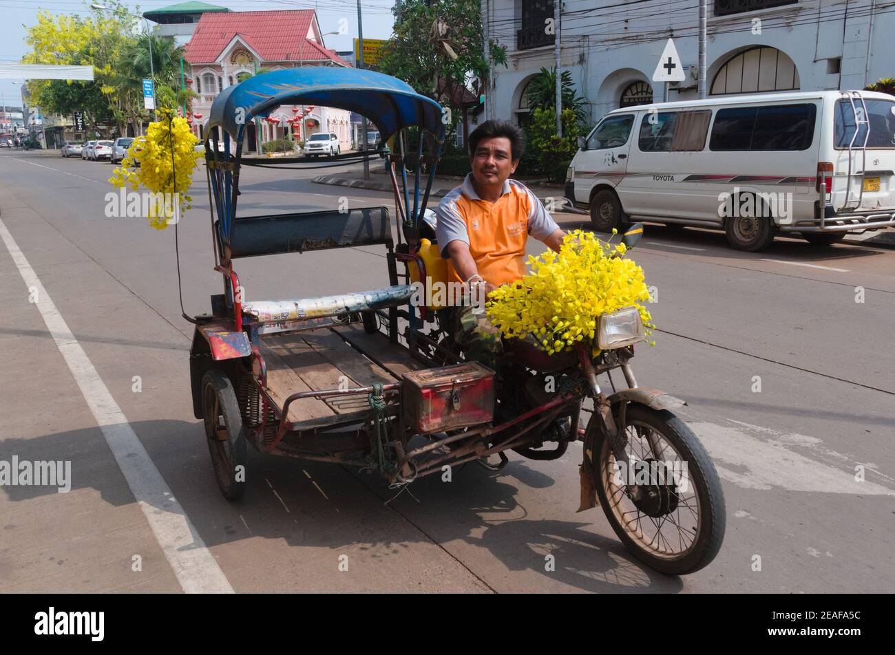 Pakse, Laos - April, 2015: Lächelnder Taxifahrer auf Dreirad-Motorrad mit Beiwagen für Passagiere geschmückt mit gelben Blumen Stockfoto