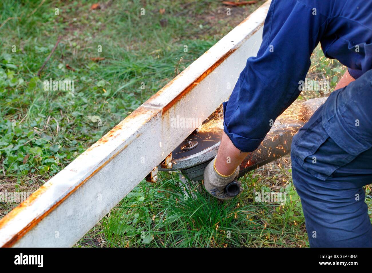 Ein Arbeiter in einem blauen Overall schneidet einen Metallbalken mit einem Winkelschleifer und schneidet Funken vor einem Hintergrund von grünem Gras. Speicherplatz kopieren. Stockfoto