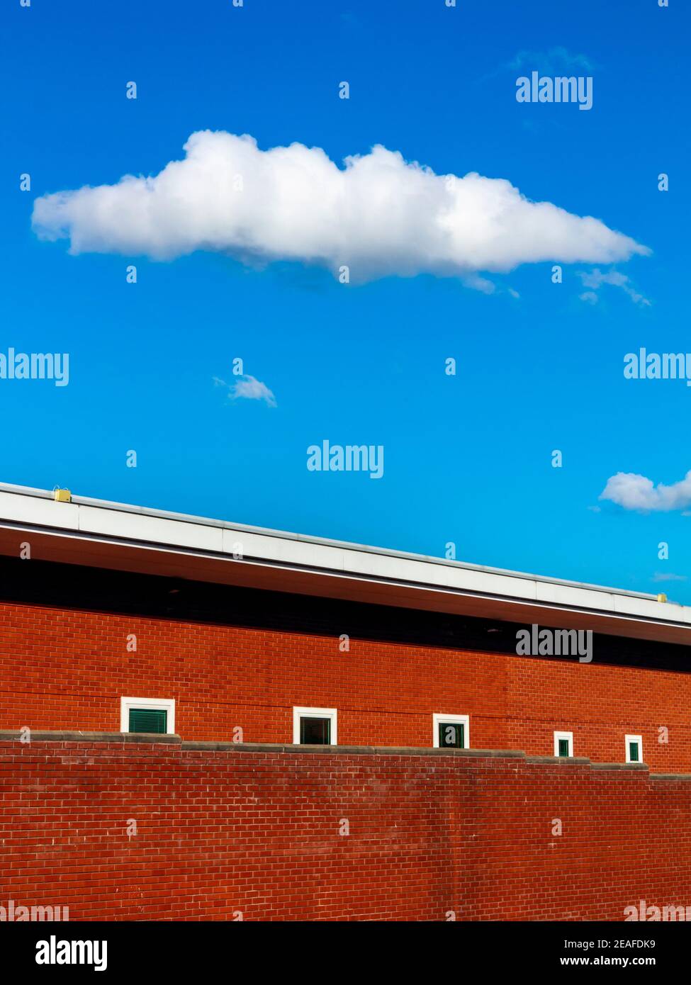Zigarrenförmige weiße Wolke in einem blauen Himmel schwebt über einem modernen roten Backsteingebäude mit kleinen Fenstern. Stockfoto