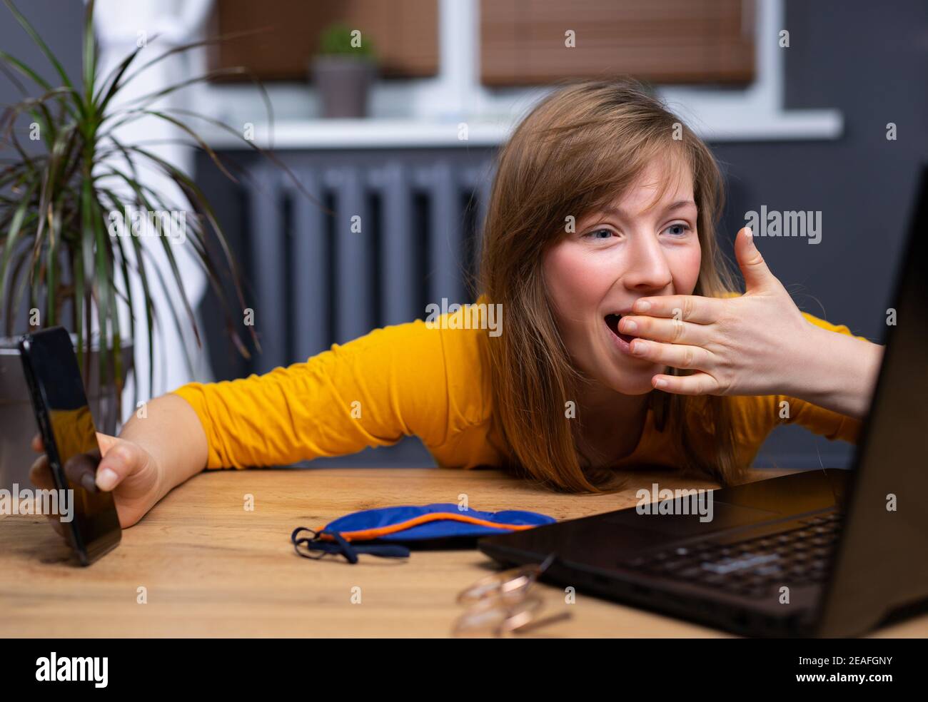 Schöne blonde Frau mit Laptop und Smartphone über Home Office Hintergrund mit Hand auf Mund. Schlafmaske auf dem Tisch, schockiert und Überraschungsgesicht Stockfoto