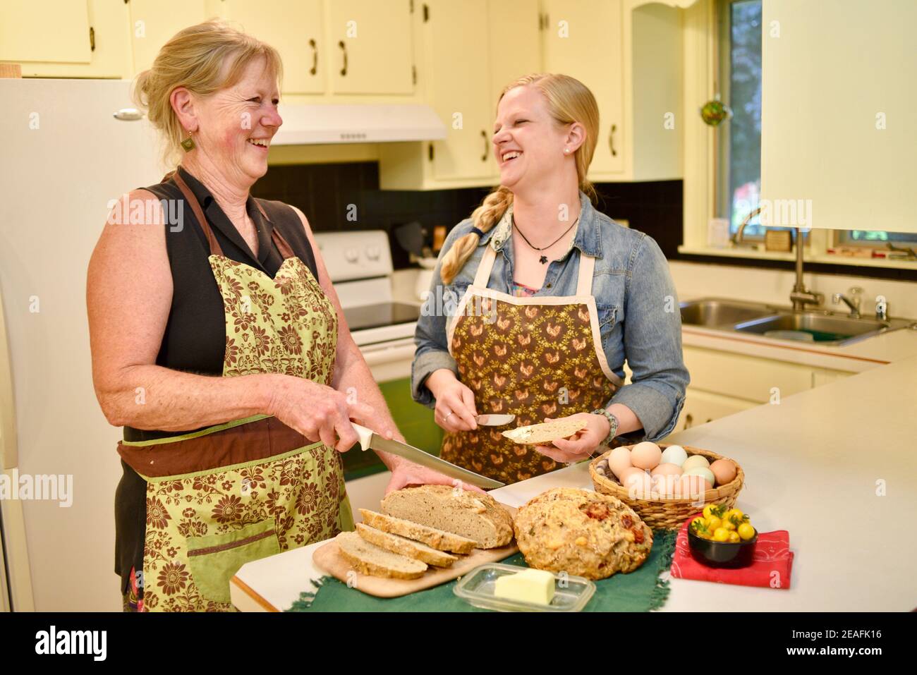 Zwei attraktive kaukasische Frauen Hausbäcker in Schürzen, schneiden frisch gebackenes Brot auf der Küchentheke, Butter Brot, Wisconsin, USA Stockfoto