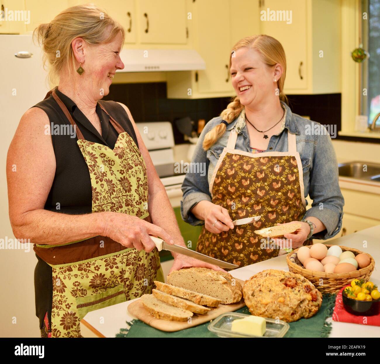 Zwei attraktive kaukasische Frauen Hausbäcker in Schürzen, schneiden frisch gebackenes Brot auf der Küchentheke, Butter Brot, Wisconsin, USA Stockfoto