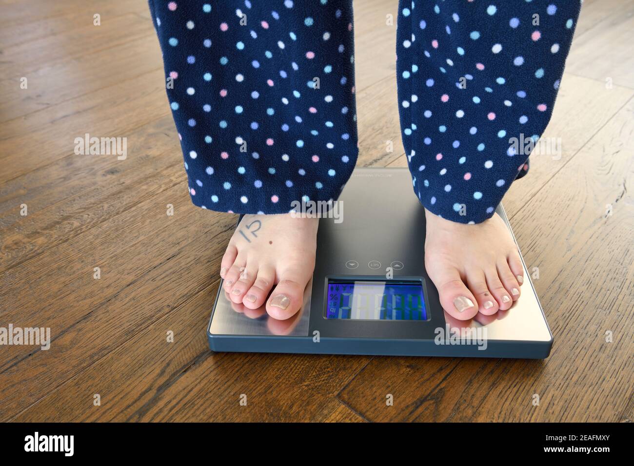 Frau im Schlafanzug und barfuß auf einer Badezimmerwaage Für Gewicht und Körperzusammensetzung auf dunklem Hartholzboden Stockfoto