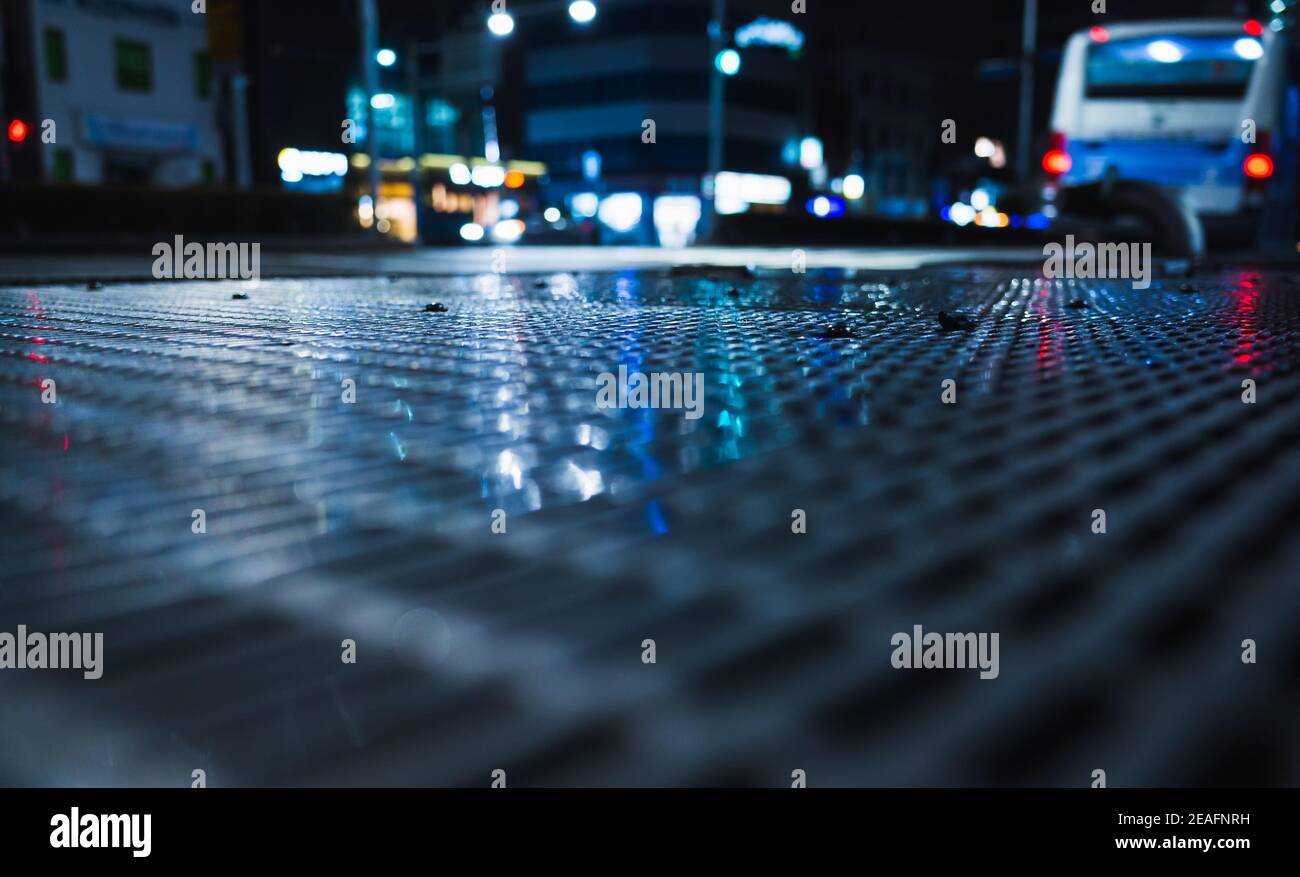 Abstrakter nächtlicher Stadthintergrund mit glänzendem nassen Straßenkanalrost und verschwommenem Licht, Foto mit selektivem Fokus Stockfoto