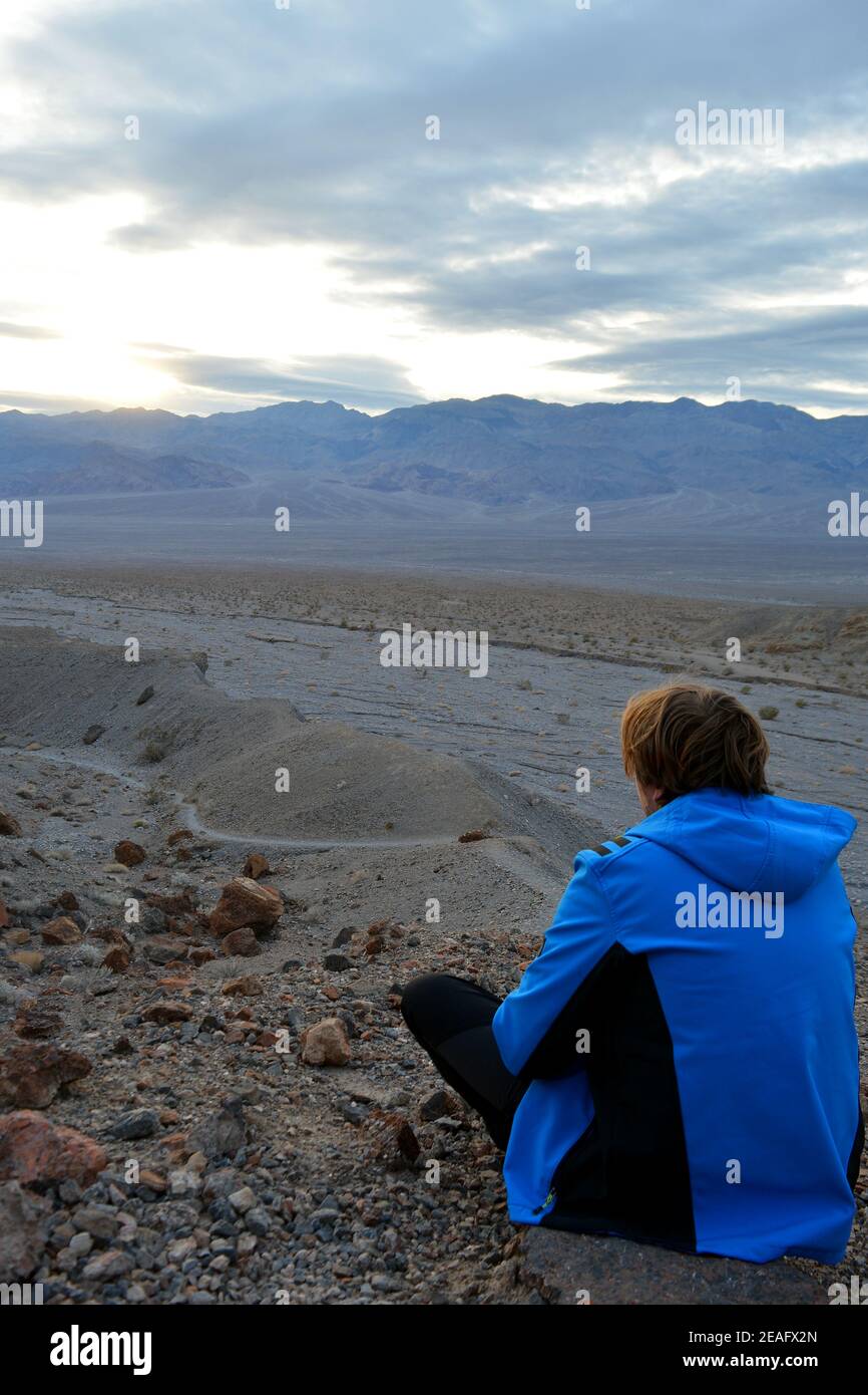 Eine Person, die auf einem Stein am Abendhimmel sitzt Mit der Sonne bricht durch Wolken mit Blick auf Die Panamint Range und das Death Valley Wash in t Stockfoto