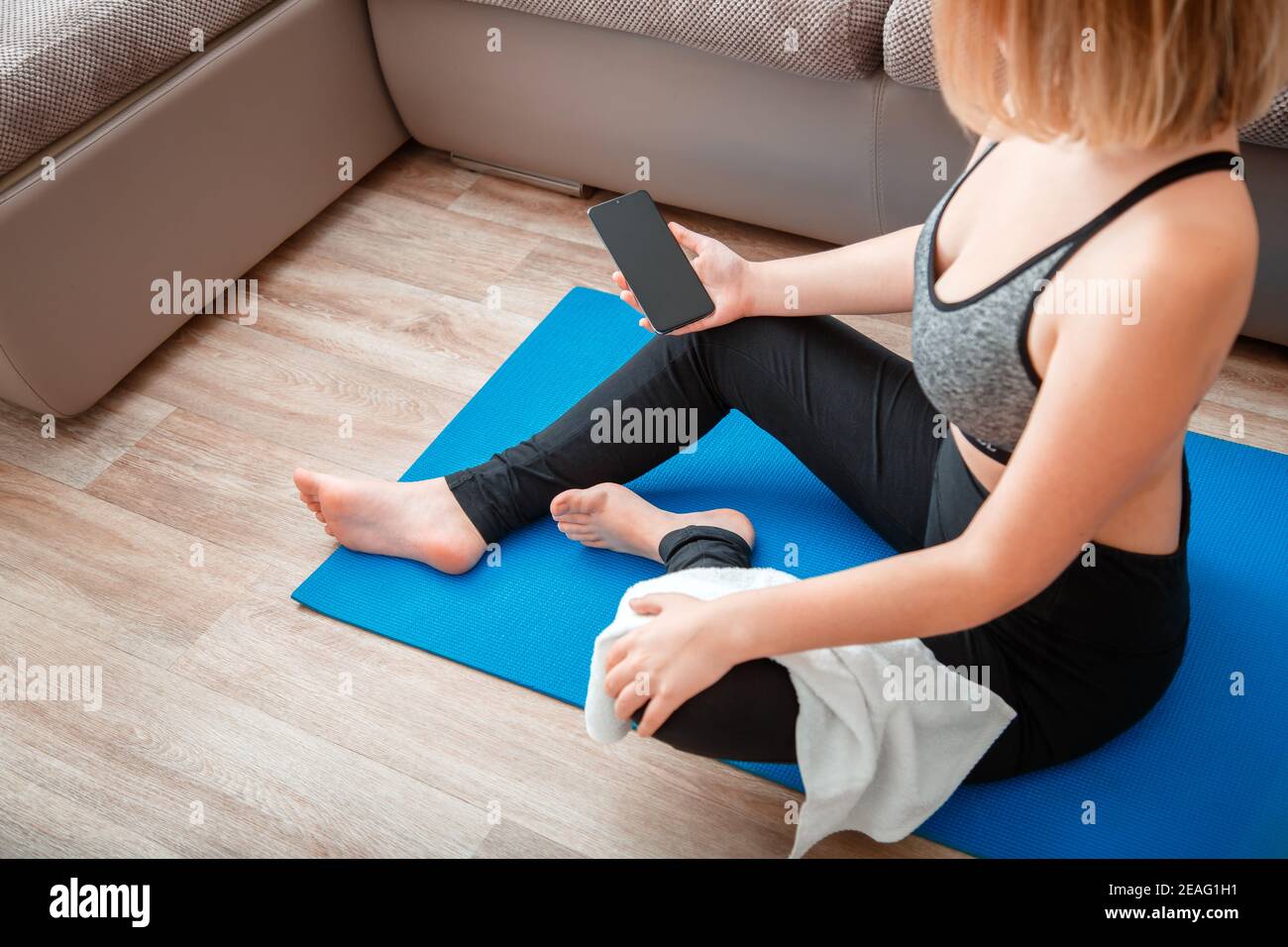 Frau in Sportbekleidung hält Smartphone mit leerem Bildschirm für Sport oder Musik-App während einer Trainingspause auf der Matte zu Hause gegen Couch. Mockup für App Stockfoto