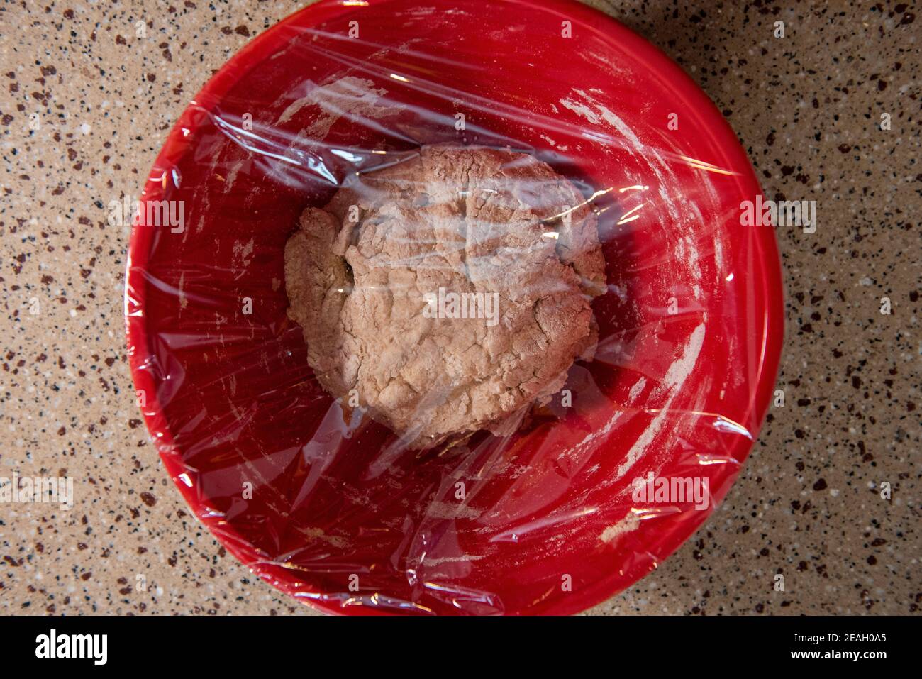 Eine Kugel Brotteig in einer roten Rührschüssel mit Plastikfolie bedeckt. Serie Schritt-für-Schritt Herstellung hausgemachtes Brot. Bild 5 von 13 Stockfoto