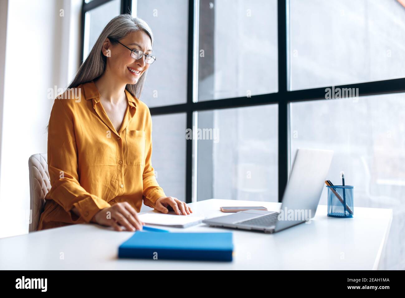 Eine reife zufriedene asiatische grauhaarige Geschäftsfrau trägt stilvolle Freizeitkleidung Brillen ist mit Laptop für Remote-Arbeit, Fernunterricht, Online-Unterricht Stockfoto