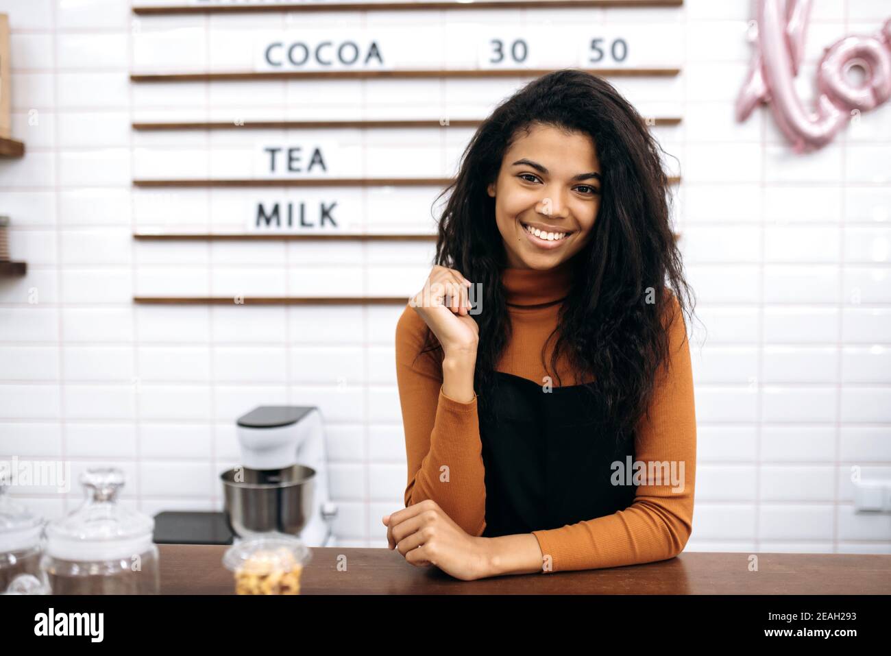 Porträt einer lächelnden hübschen Kellnerin in Uniform. Eine junge, schöne afroamerikanische Barista lächelt vor der Kamera und steht hinter der Theke im Café. Inhaber kleiner Unternehmen Stockfoto