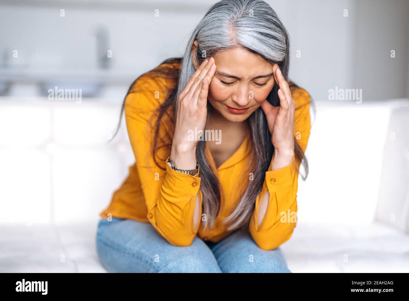Mittlere Alter grauhaarige asiatische Frau sitzt auf Couch im Wohnzimmer hält ihren Kopf mit ihren Händen, fühlt sich unglücklich wegen Kopfschmerzen, persönliche Probleme, Krankheit oder schlechte Nachrichten, sie brauchen psychologische oder Stockfoto