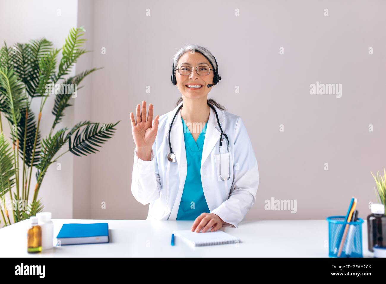 Die grauhaarige Ärztin im mittleren Alter trägt ein Headset, schaut auf die Kamera und lächelt. Remote-Online-medizinische Beratung, Medizin Ferndienste, virtuelle medizinische Hilfe Konzept Stockfoto