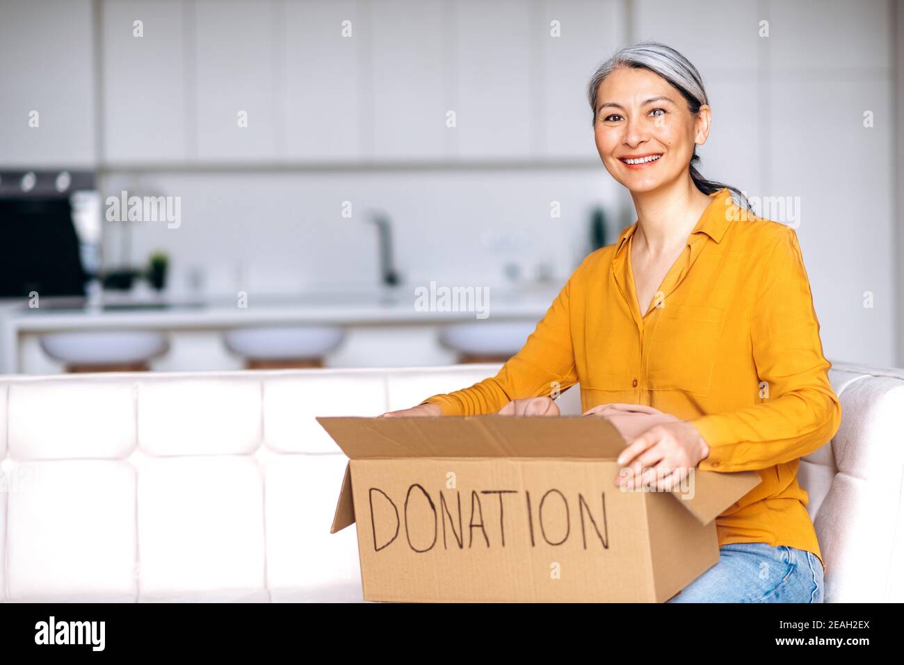 Spendenkonzept. Eine zufriedene Frau mittleren Alters mit grauem Haar, die zu Hause auf dem Sofa sitzt, hält eine Spendenbox mit voller Kleidung für die Wiederverwendung oder das Recycling alter Kleidung. Wohltätigkeitsorganisation. Hilfe für arme und bedürftige Menschen Stockfoto