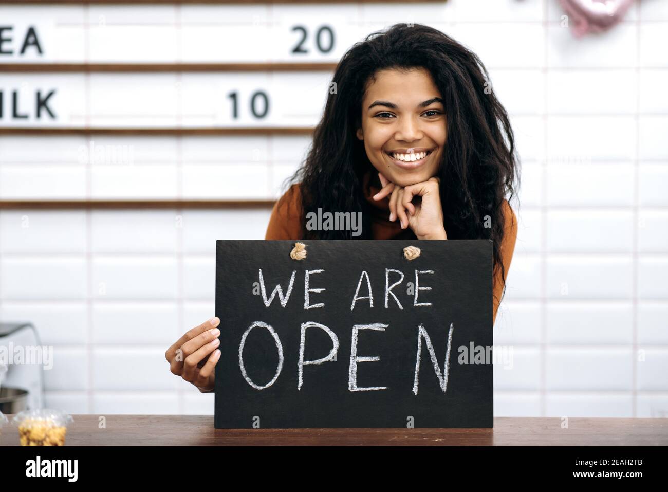 Herzlich Willkommen. Porträt einer glücklichen afroamerikanischen Barista oder Kellnerin, Besitzer eines Coffee-Shop, steht mit einem Schild, das wir hinter der Bar-Theke öffnen, freundlich lächelnd und wartet auf Besucher Stockfoto