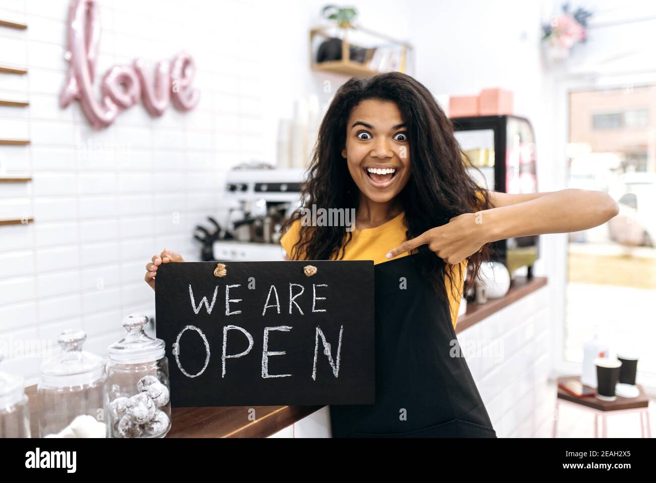 Aufgeregte afroamerikanische Barista oder Kellnerin, Inhaberin eines Coffee Shops oder Restaurants freut sich über die Eröffnung, hält ein Schild, DAS WIR IN der Nähe der Theke ÖFFNEN und überglücklich lächelt Stockfoto