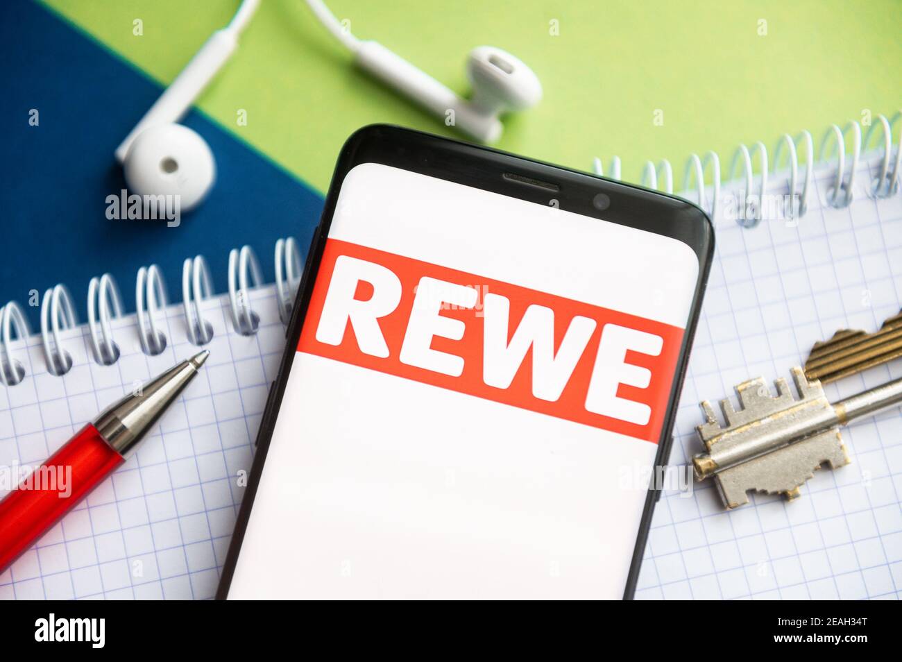 In dieser Abbildung wird ein Rewe-Logo auf einem Smartphone mit Stift,  Schlüssel, Buch und Headsets im Hintergrund angezeigt Stockfotografie -  Alamy
