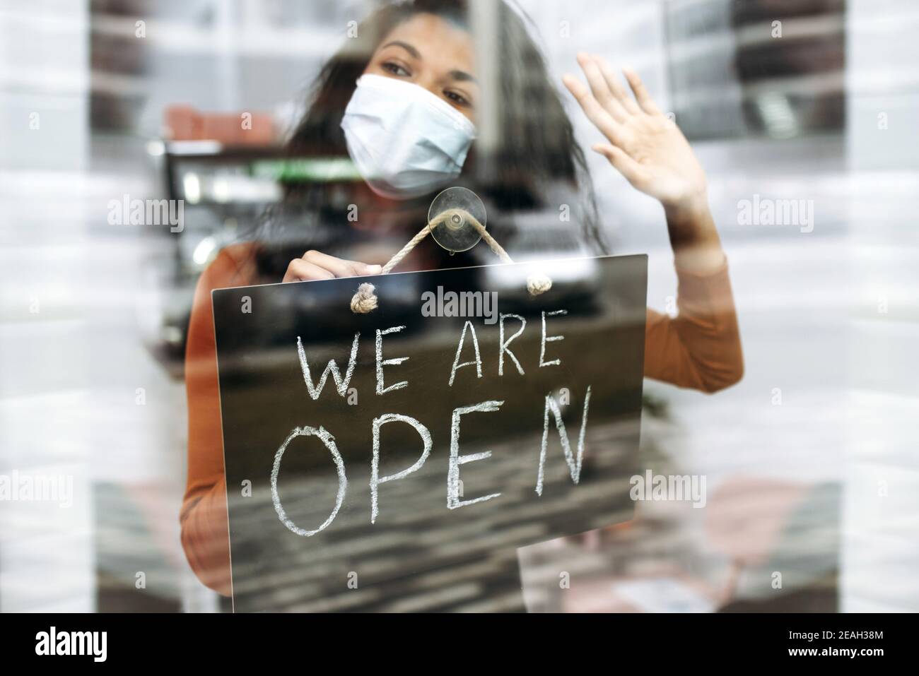 Afroamerikanische Kleinunternehmerin in medizinische Maske hält ein Schild, DAS WIR am Eingang zu einem Restaurant, Bar, Café oder Shop öffnen, und wartet auf Besucher Stockfoto