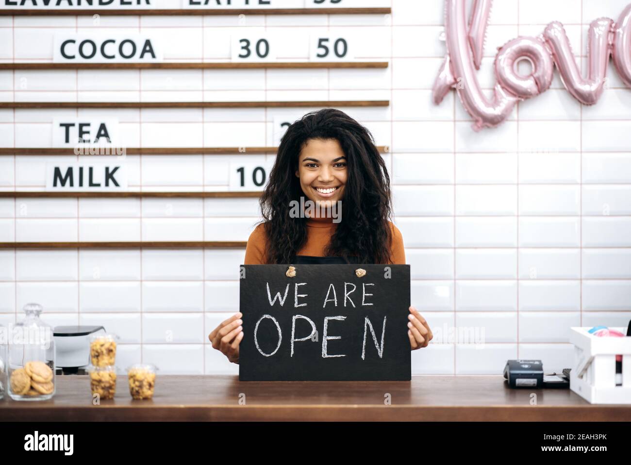 Glückliche afroamerikanische Barista oder Kellnerin, Inhaberin eines Coffee-Shops, steht mit einem Schild, das wir hinter der Theke öffnen und freundlich lächeln Stockfoto