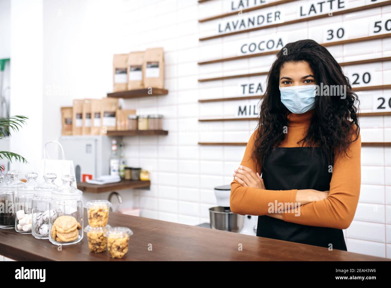 Porträt einer hübschen Kellnerin in Uniform und schützende medizinische Maske. Die junge afroamerikanische Barista steht hinter der Theke und schaut auf die Kamera. Konzept für kleine Unternehmen Stockfoto