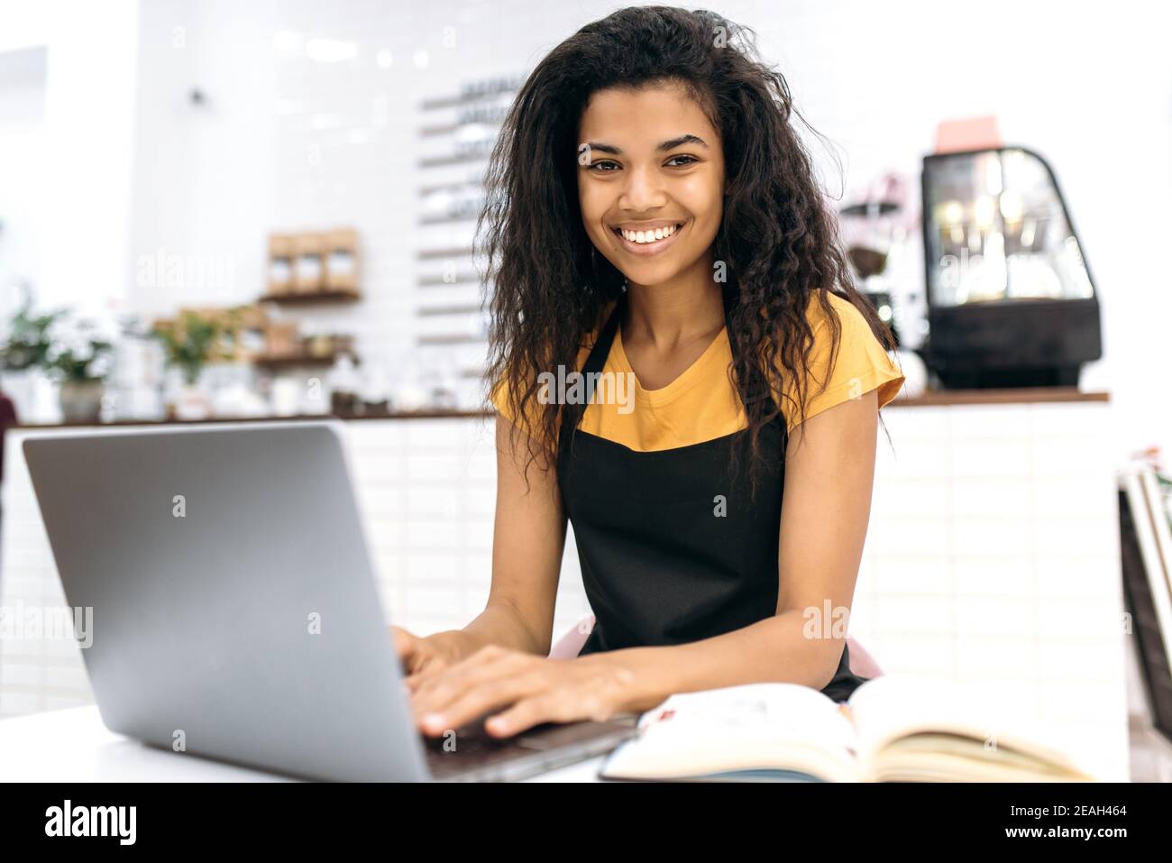 Zufriedene junge afroamerikanische weibliche Kleinunternehmerin oder Kellnerin, studiert Kleinunternehmen monatliche Abrechnung mit Laptop, schaut auf die Kamera mit Lächeln, sitzen in ihrem Café Stockfoto