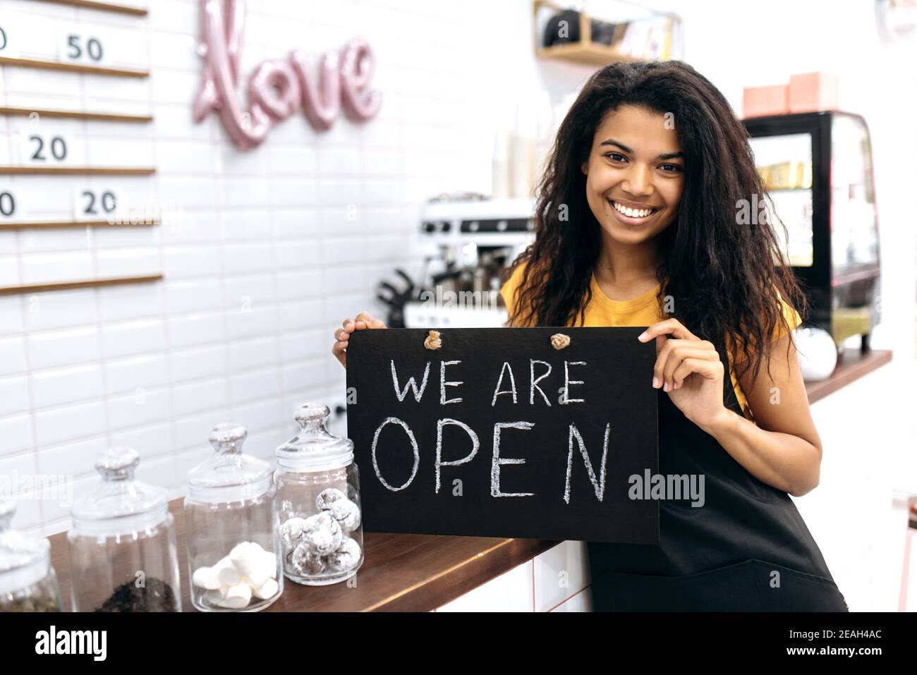 Zufriedene afroamerikanische Barista oder Kellnerin, Inhaberin eines Kaffeehauses oder Restaurants, steht mit einem Schild, das wir in der Nähe der Theke öffnen und freundlich lächeln Stockfoto