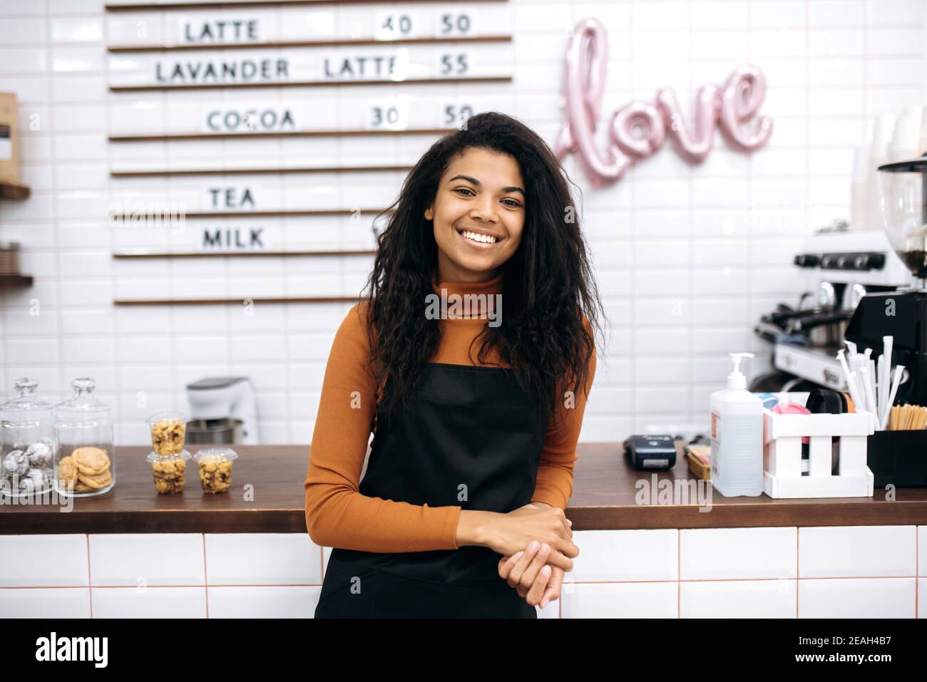 Kaffee-Business-Eigentümer Konzept. Attraktive junge afroamerikanische Barista oder Kellnerin in schwarzer Schürze freundlich lächelt an der Kamera in der Nähe von Café-Theke Bar. Stockfoto