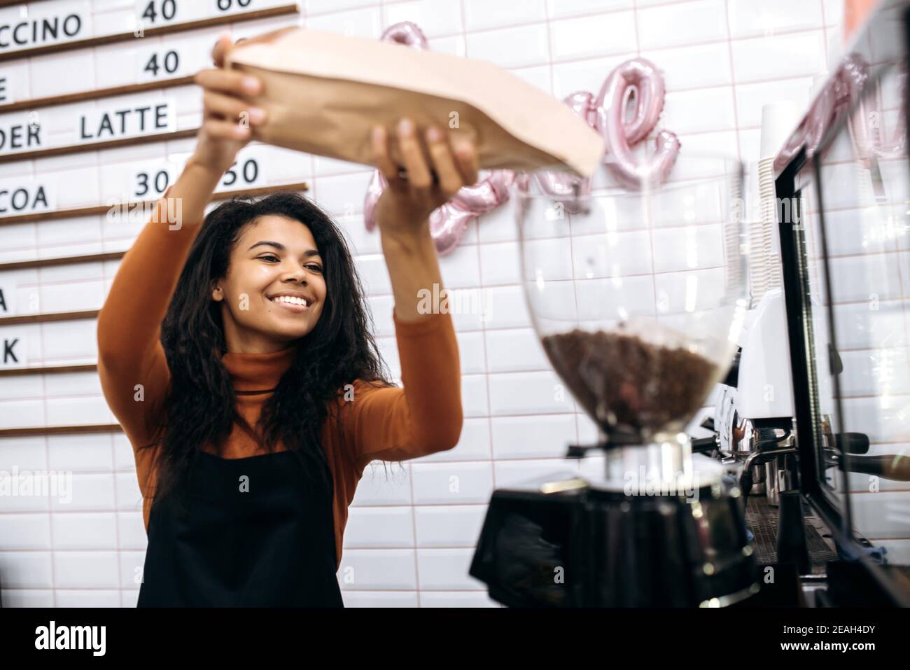 Eine zufriedene afroamerikanische Barista in einer schwarzen Schürze gießt aromatische Kaffeebohnen in eine Kaffeemaschine. Konzept des Inhabers kleiner Unternehmen Stockfoto