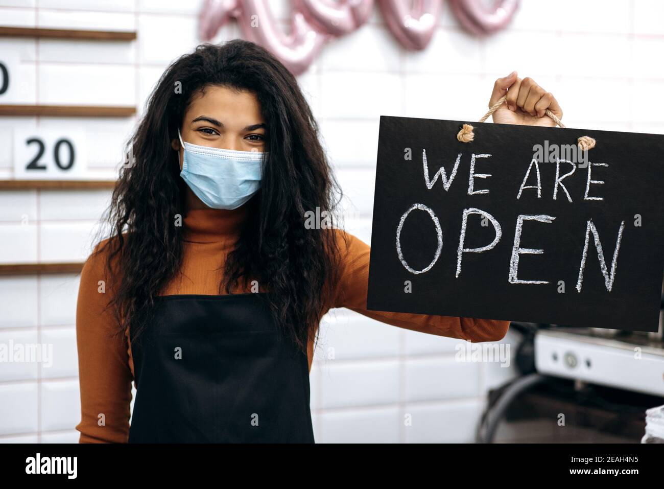 Afroamerikanische Barista oder Kellnerin, oder Besitzer eines Coffee-Shop, trägt eine medizinische Maske, steht mit einem Schild, DAS WIR IN einem Café ÖFFNEN und warten auf Besucher Stockfoto
