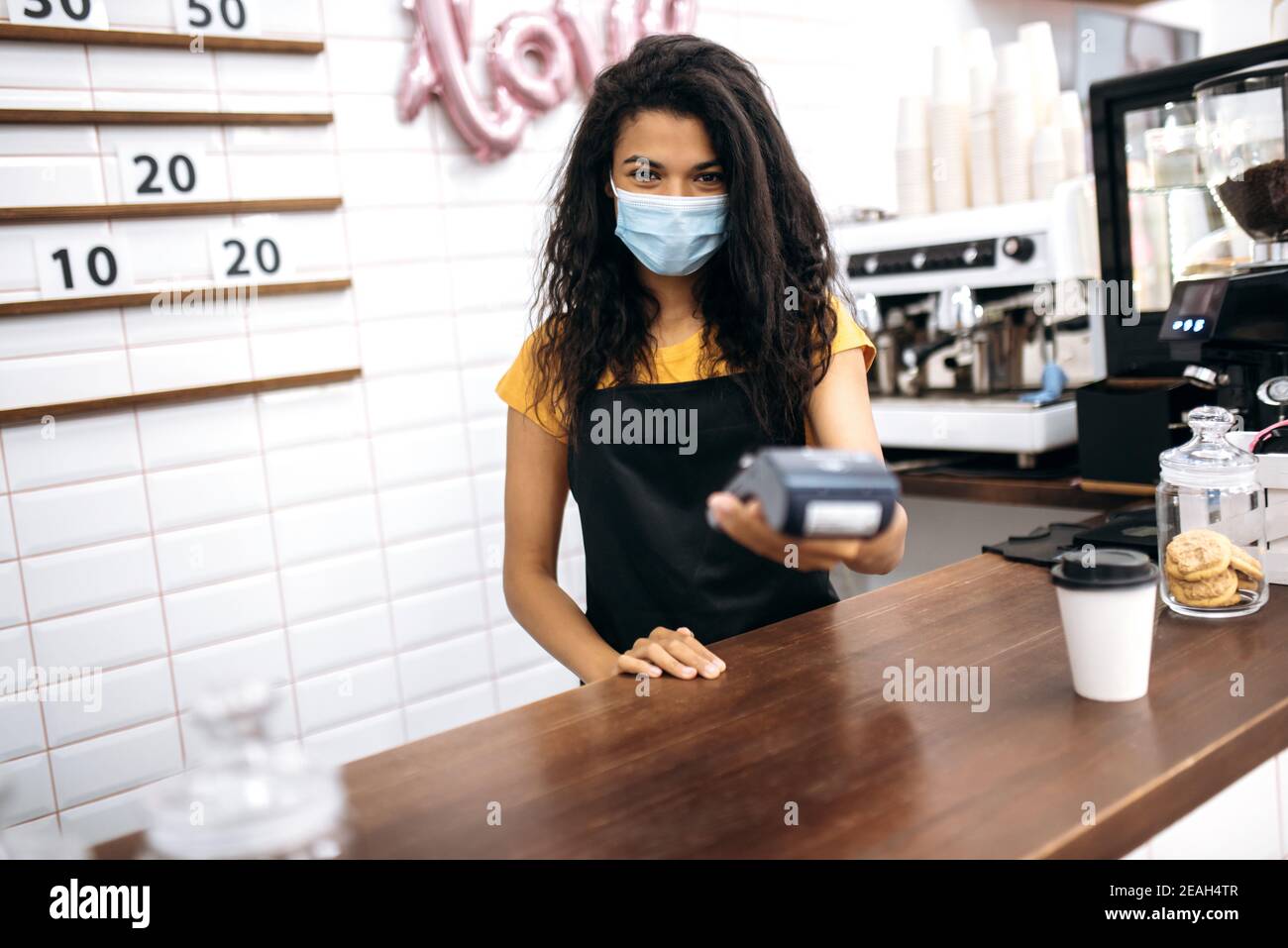 afroamerikanische Mädchen Barista, Kellnerin oder Kleinunternehmen Inhaber trägt schützende medizinische Maske geben Besucher ein kontaktloses Zahlungsterminal Stockfoto