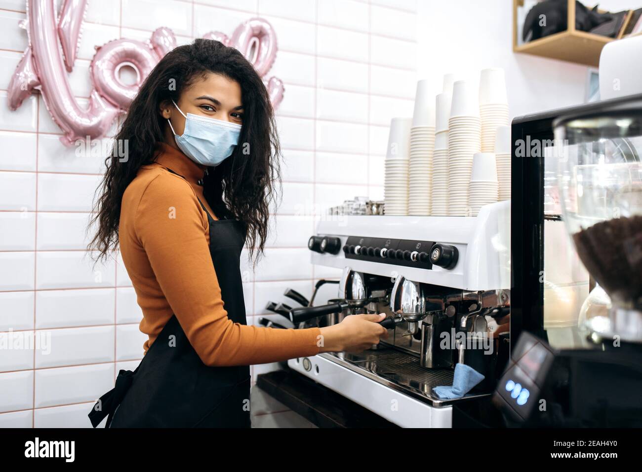 Die junge afroamerikanische Barista trägt eine medizinische Maske und eine schwarze Schürze und macht Kaffee für einen Café-Besucher bei ihnen. Inhaber eines kleinen Unternehmens. Café, Service-Industrie-Konzept Stockfoto
