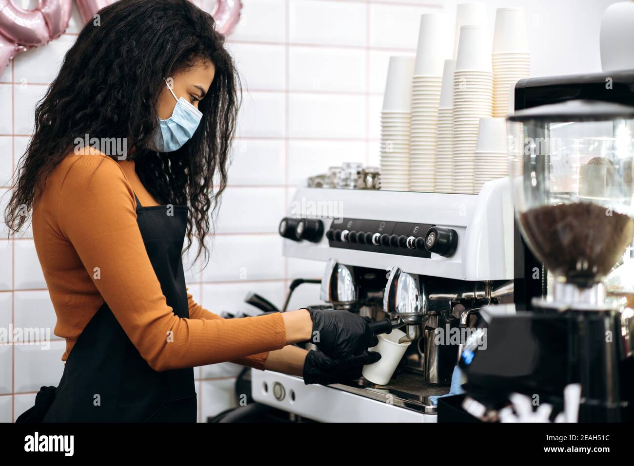Die junge afroamerikanische Barista trägt eine medizinische Maske und eine schwarze Schürze und macht Kaffee für einen Café-Besucher bei ihnen. Inhaber eines kleinen Unternehmens. Café, Service-Industrie-Konzept Stockfoto