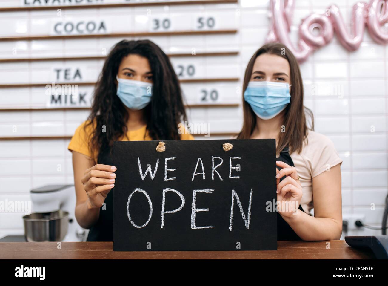 Zwei entkochte weibliche Kellnerin ein Café oder Restaurant, afroamerikanisch und kaukasisch, steht mit einem Schild, DAS WIR HINTER der Theke mit medizinischen Masken ÖFFNEN Stockfoto