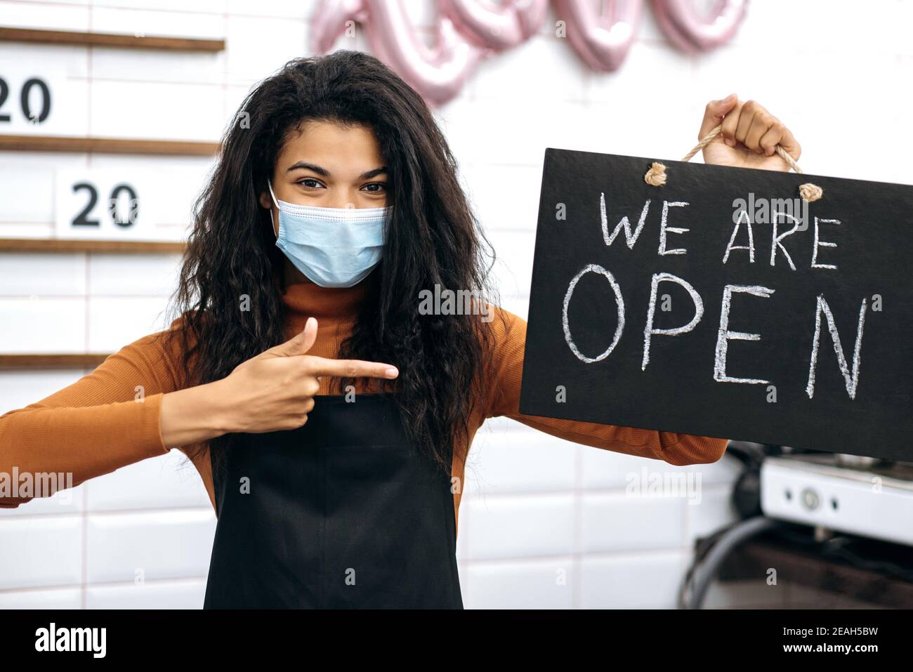 Multirassische lockige weibliche Barista oder Kellnerin eines Coffee-Shops, trägt eine medizinische Maske, hält und Punkte auf einem Schild SIND WIR IN einem Café öffnen und wartet auf Besucher Stockfoto