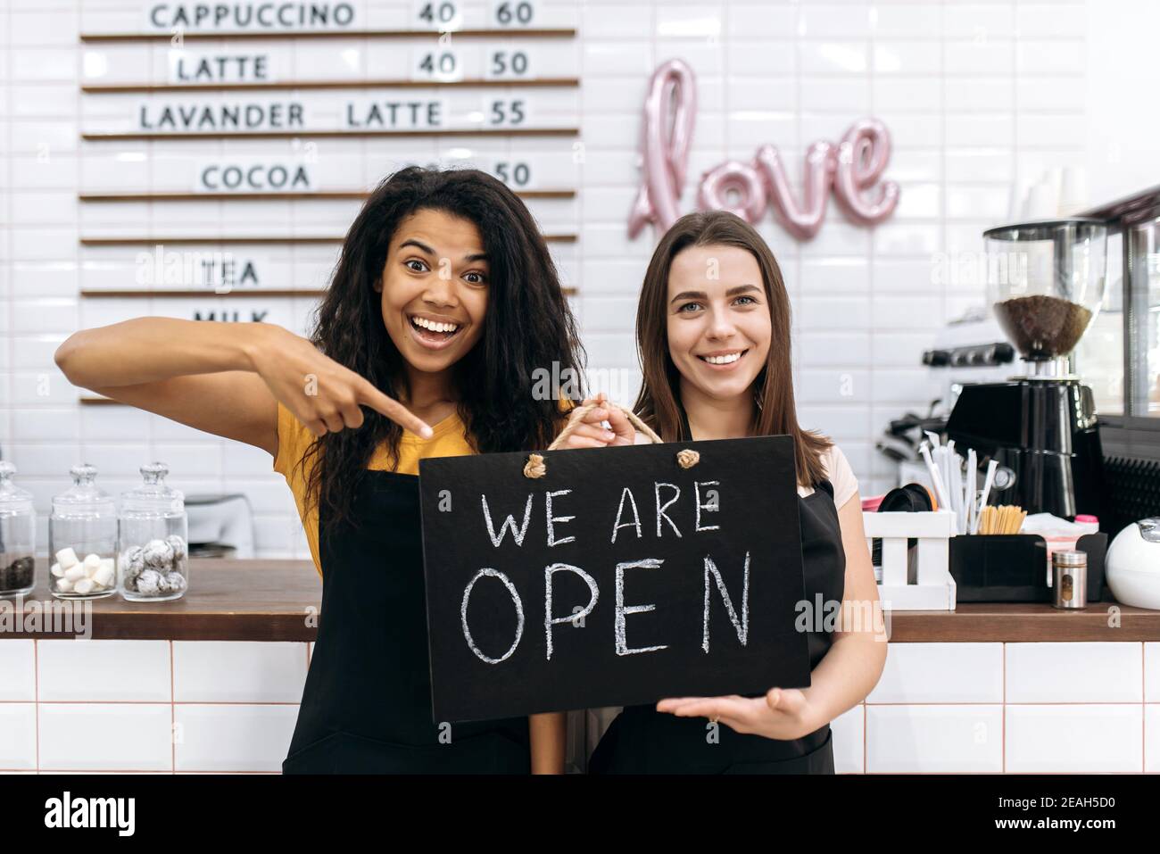 Zwei zufriedene weibliche Kellnerin ein Café oder Restaurant, afroamerikanisch und kaukasisch, steht mit einem Schild, DAS WIR IN der Nähe der Theke öffnen und freundlich lächeln Stockfoto