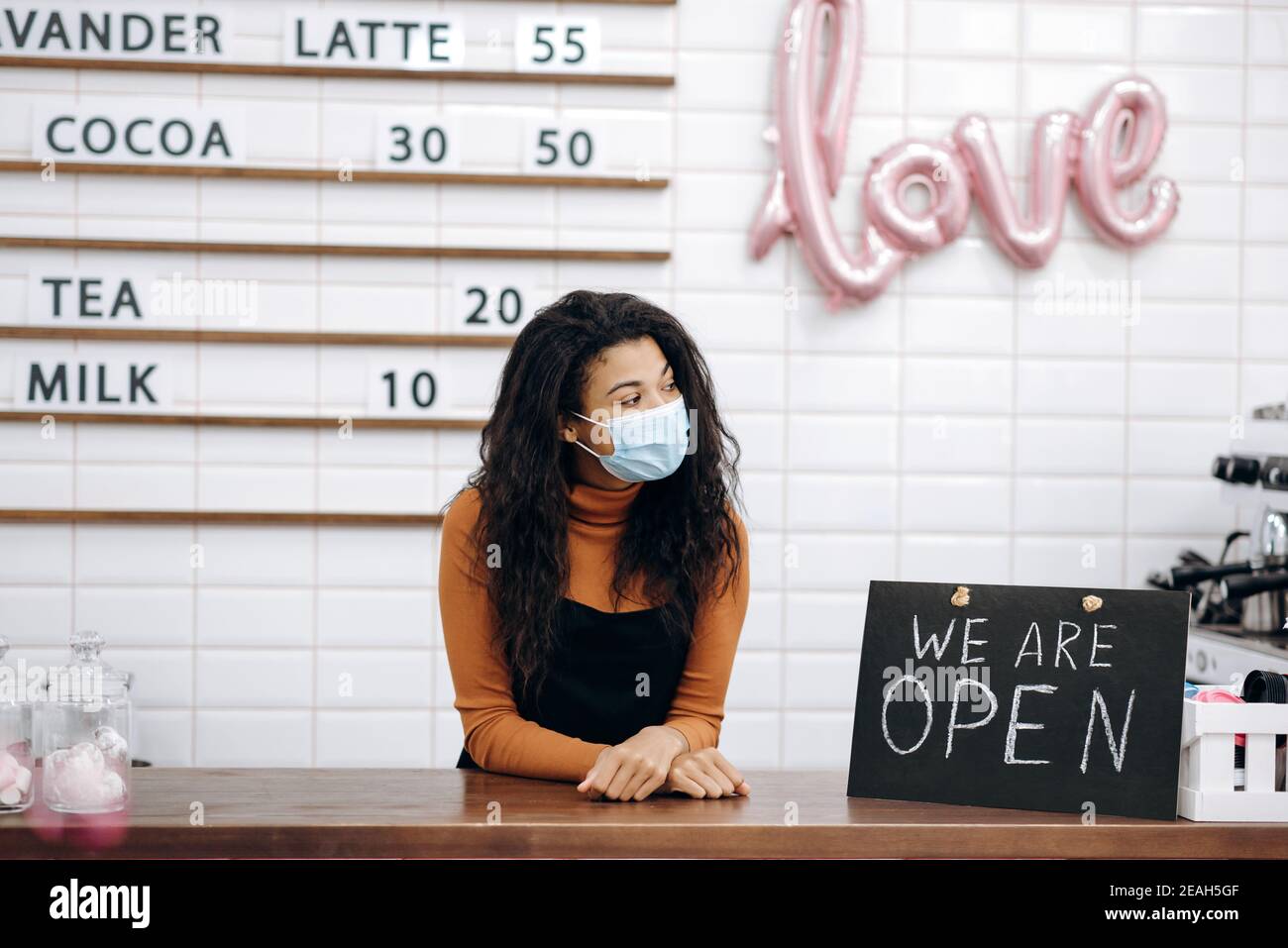 Eine freundliche afroamerikanische Barista, Kellnerin oder Cafébesitzerin, die eine medizinische Maske trägt, steht hinter der Theke neben einem Schild, DAS WIR ÖFFNEN, und wartet auf Besucher Stockfoto