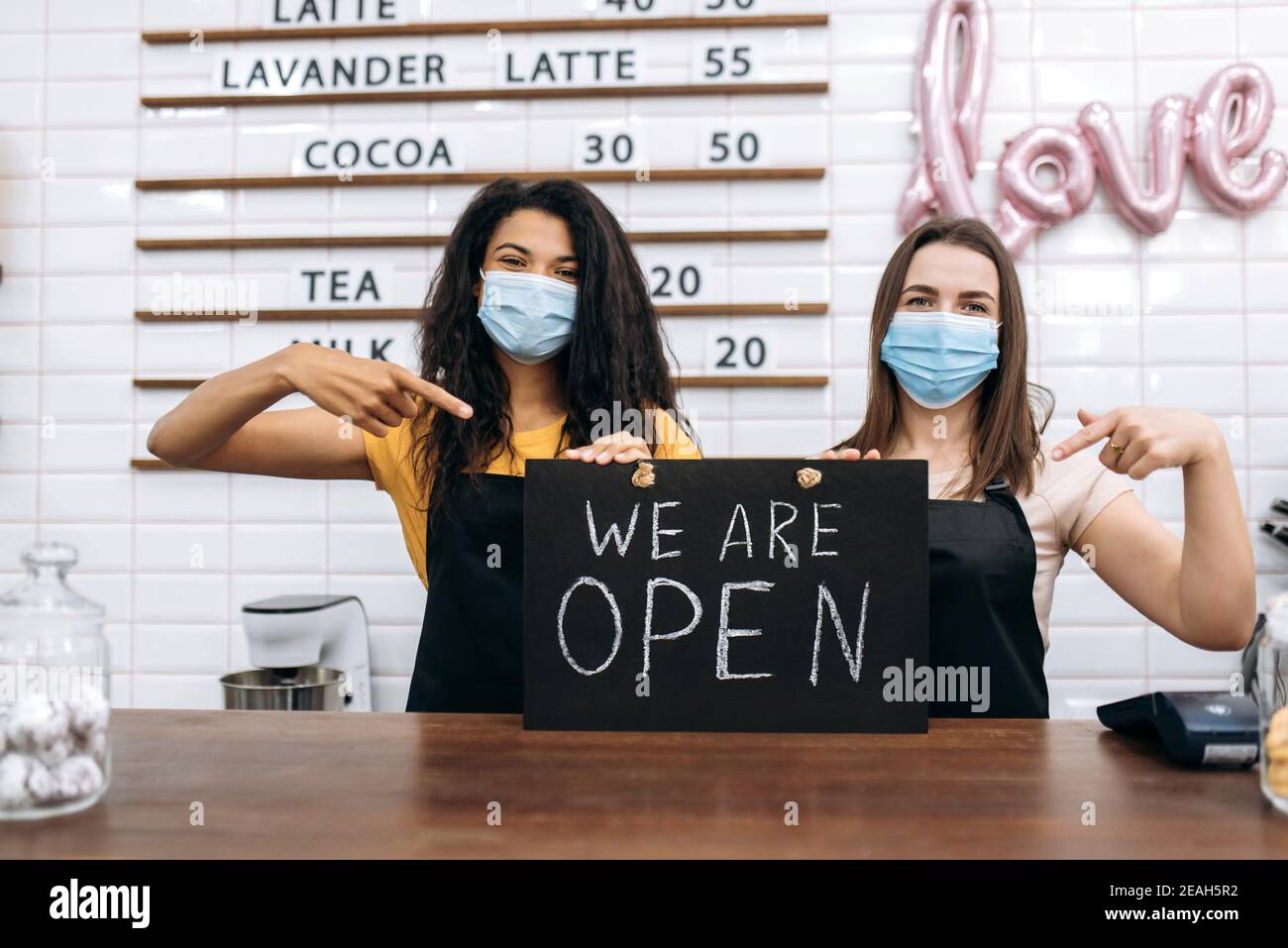 Zwei zufriedene weibliche Kellnerin ein Café oder Restaurant, afroamerikanisch und kaukasisch, steht mit einem Schild, DAS WIR HINTER der Theke mit medizinischen Masken ÖFFNEN Stockfoto