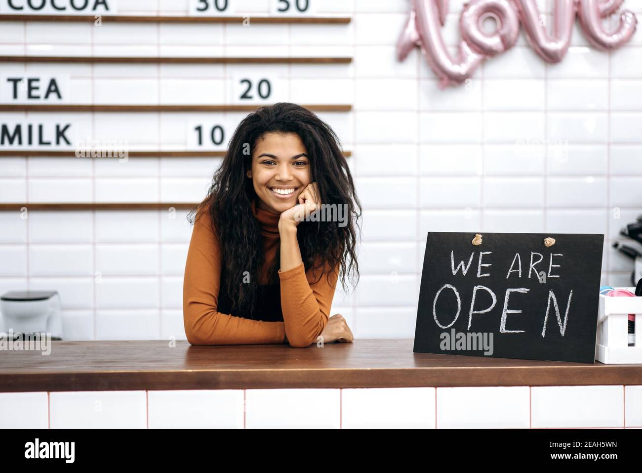 Herzlich Willkommen. Glückliche afroamerikanische Barista oder Kellnerin, Inhaberin eines Coffee-Shops, steht mit einem Schild, DAS WIR HINTER der Theke öffnen und freundlich lächeln Stockfoto