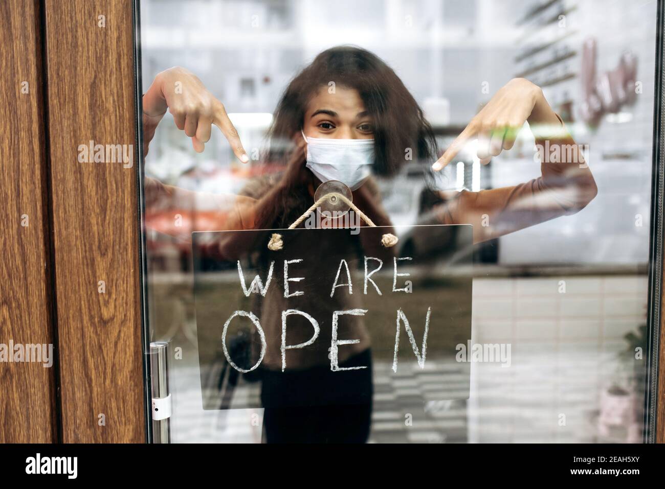 Lockige weibliche afroamerikanische Kleinunternehmerin in medizinische Maske Punkte an einem Schild SIND WIR AM Eingang zu einem Restaurant, Bar, Café oder Shop geöffnet, und wartet auf Besucher Stockfoto