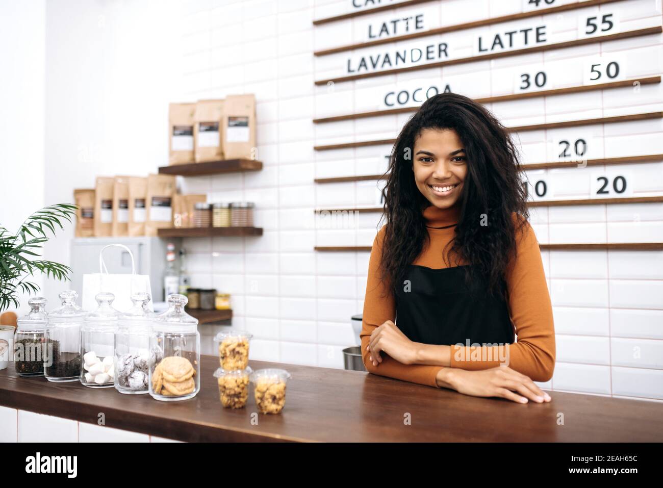 Freundliche, hübsche Kellnerin in Uniform. Eine junge, lockige, afroamerikanische Barista lächelt vor der Kamera und steht hinter der Theke im Café. Konzept für kleine Unternehmen Stockfoto