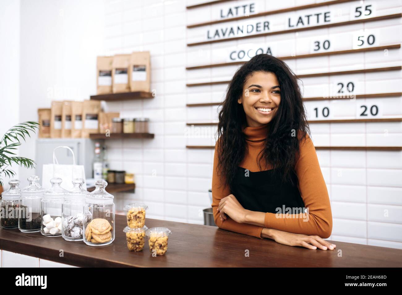 Glücklich zufrieden Kellnerin trägt Uniform. Eine junge, lockige, afroamerikanische Barista lächelt vor der Kamera und steht hinter der Theke im Café. Konzept für kleine Unternehmen Stockfoto