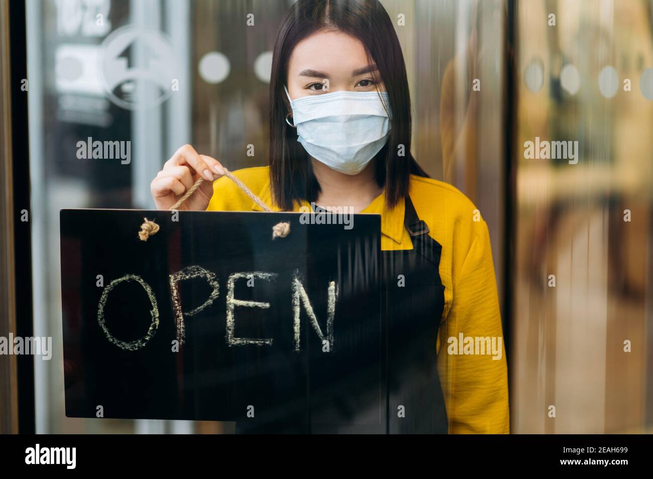 Porträt einer glücklichen Kellnerin, die am Eingang des Restaurants oder Cafés steht und eine medizinische Maske auf ihrem Gesicht trägt. Junge asiatische Frau zeigt offene Zeichen in ihrem kleinen Geschäft und lädt Kunden Stockfoto