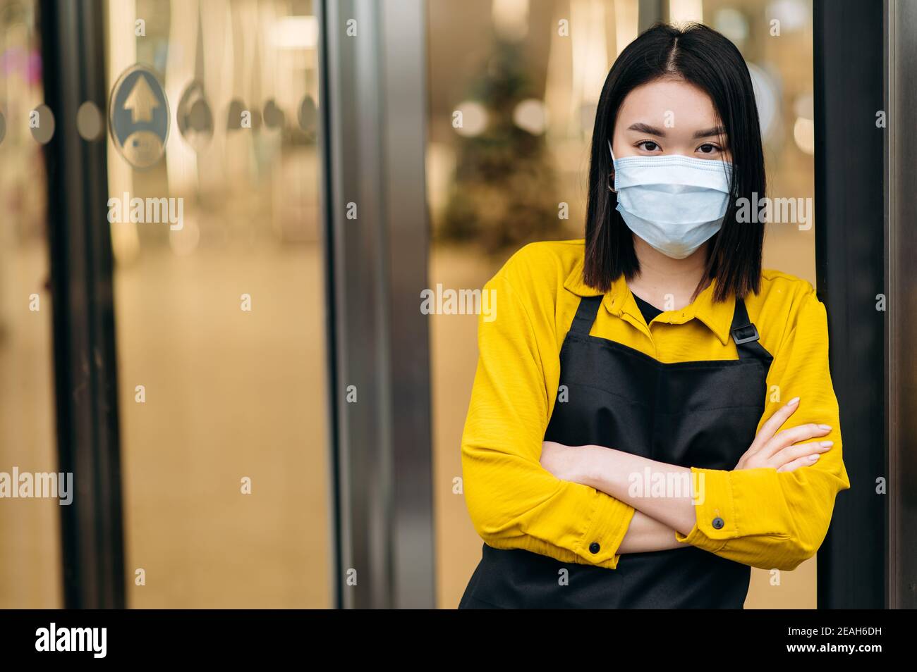 Eine junge asiatische Kellnerin oder Inhaberin eines Restaurants, Cafés oder Shops in einer Schutzmaske und einer Schürze steht am Eingang. Kleinunternehmen inmitten der Coronavirus-Pandemie Stockfoto