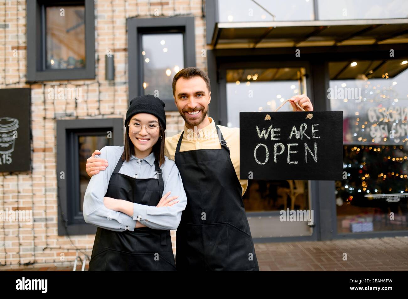 Wir sind OFFEN. Zwei multirassische freundliche Kellner in Uniform stehen vor einem Restaurant, Café oder Bar, zeigen Schild OFFEN, mit einem angenehmen Lächeln, Herzlich Willkommen. Teamwork-Konzept Stockfoto