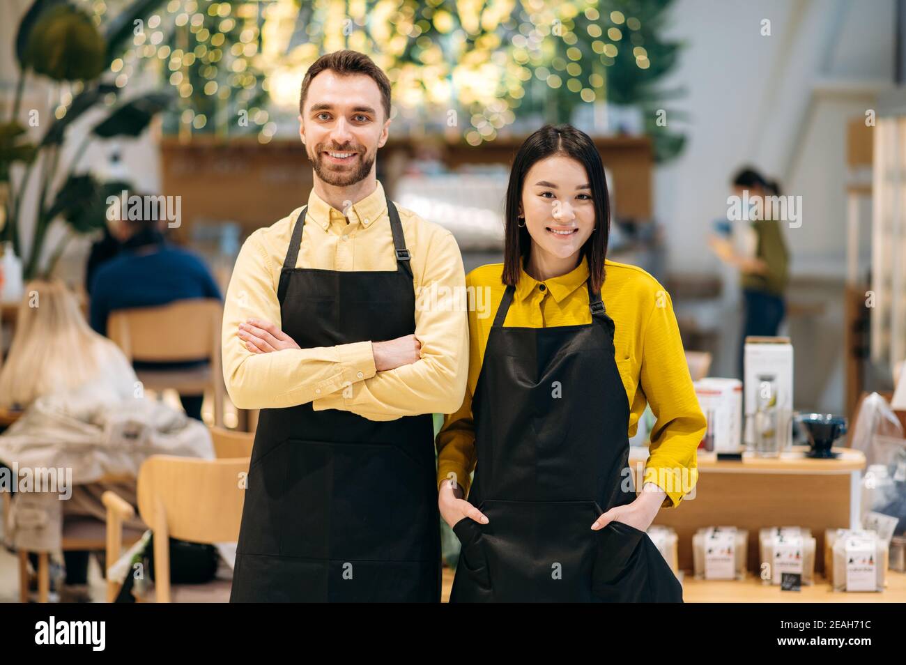 Teamarbeit. Porträt von zwei freundlichen Kellnern. Attraktive kaukasischen Kerl und schöne asiatische Mädchen in schwarzen Schürzen stehen in einem Restaurant, Café oder Bar und lächelnd Stockfoto