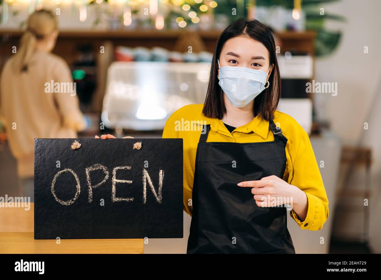 Schild GEÖFFNET. Porträt einer jungen asiatischen Kellnerin, die in einem Restaurant oder Café mit Schutzmaske und schwarzer Schürze steht und das Schild offen hält. Unterstützung des Konzepts für kleine Unternehmen Stockfoto