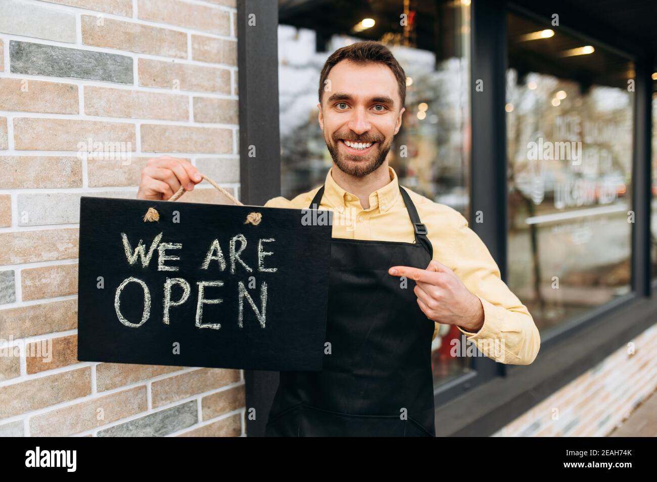 Willkommen, wir sind offen. Porträt des kaukasischen attraktiven männlichen Kellner im Freien in der Nähe eines Restaurants oder Cafés, zeigt einen Finger auf ein Schild OFFEN und freundliches Lächeln. Unterstützung des Konzepts für kleine Unternehmen Stockfoto